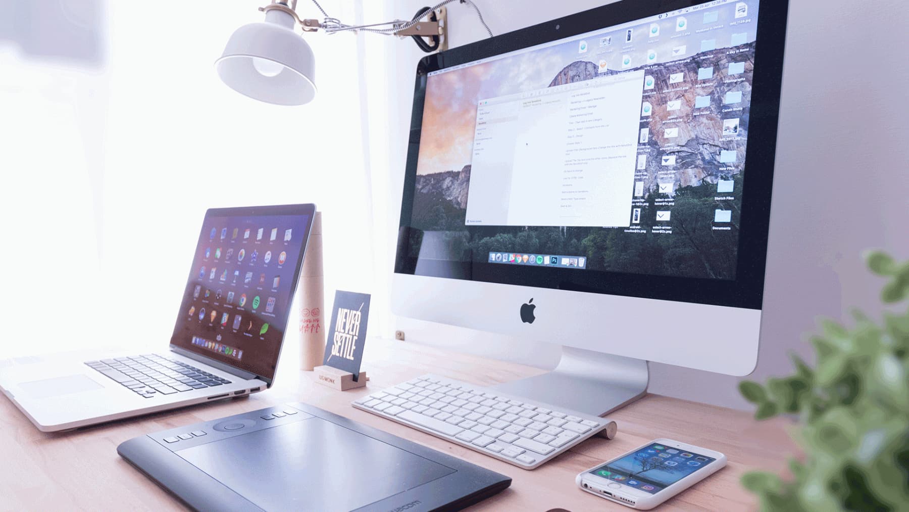 Apple MacBook and iMac displaying web browser content, with keyboard, mouse and Never Settle block on wooden desk